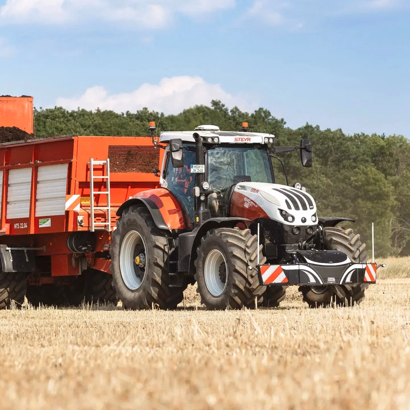 Red and white tractor with a front weight bumper attached, pulling a large red trailer in a harvested field