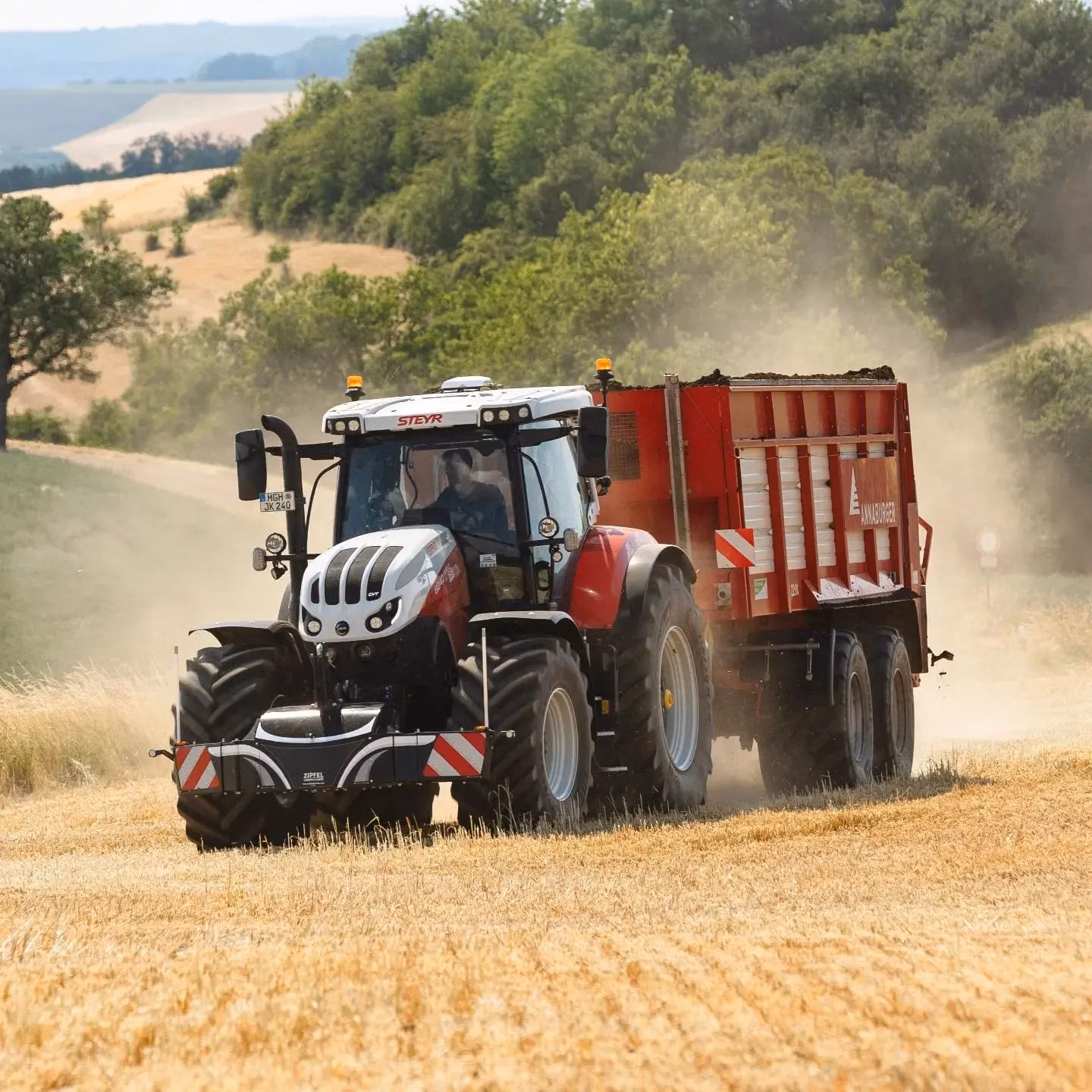 A tractor with a front weight bumper and large trailer drives through a dry, harvested field with dust in the air