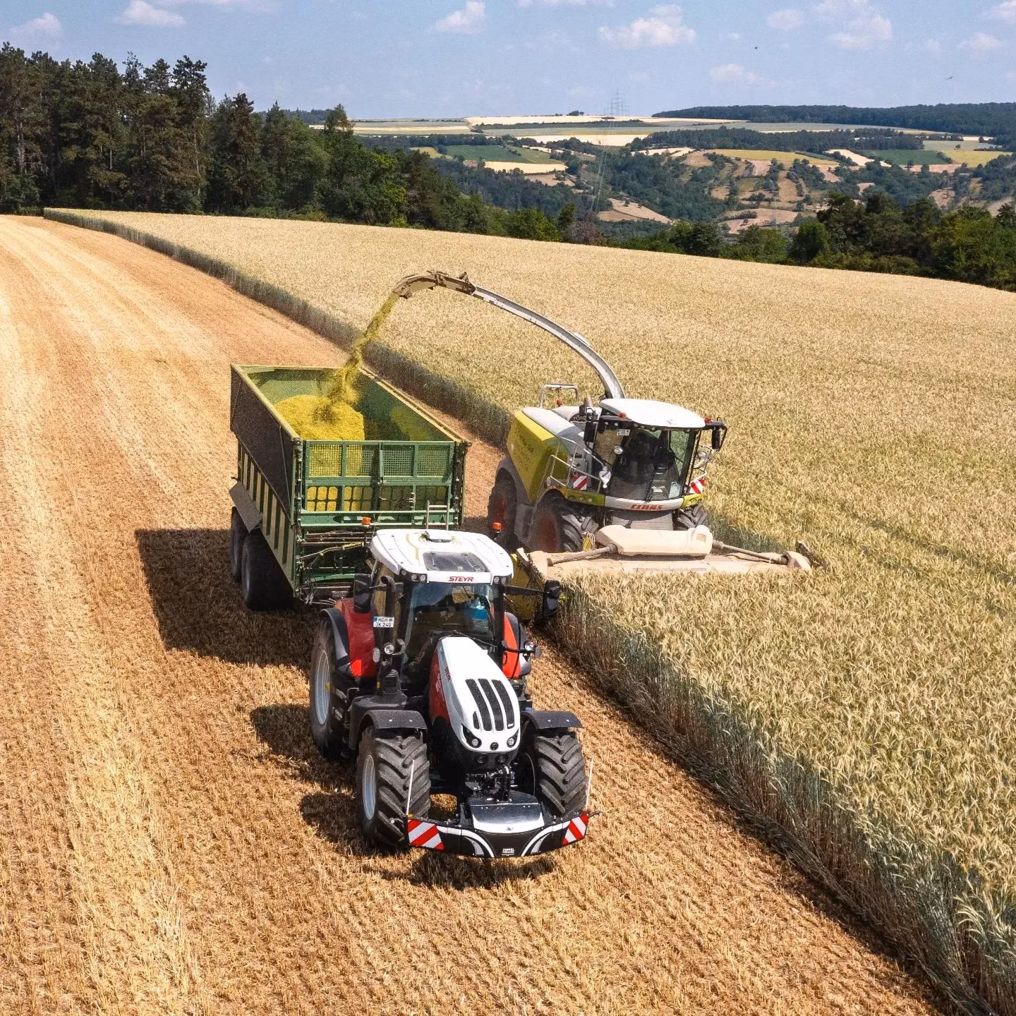 A tractor with a front weight bumper pulls a trailer in a field beside a forage harvester unloading crops