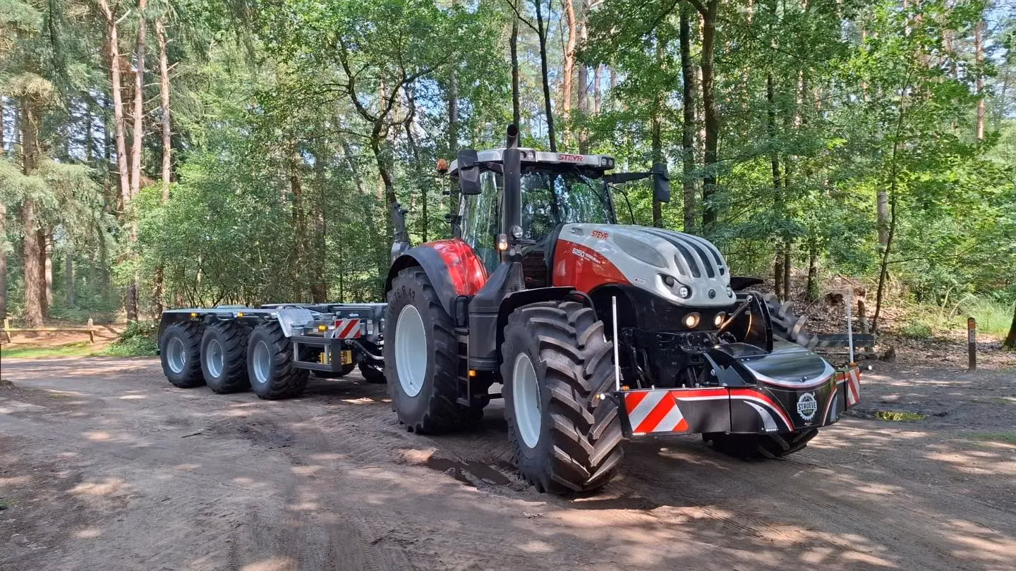 Red and silver tractor with a front weight bumper and LED lighting, towing a multi-axle trailer on a forest path
