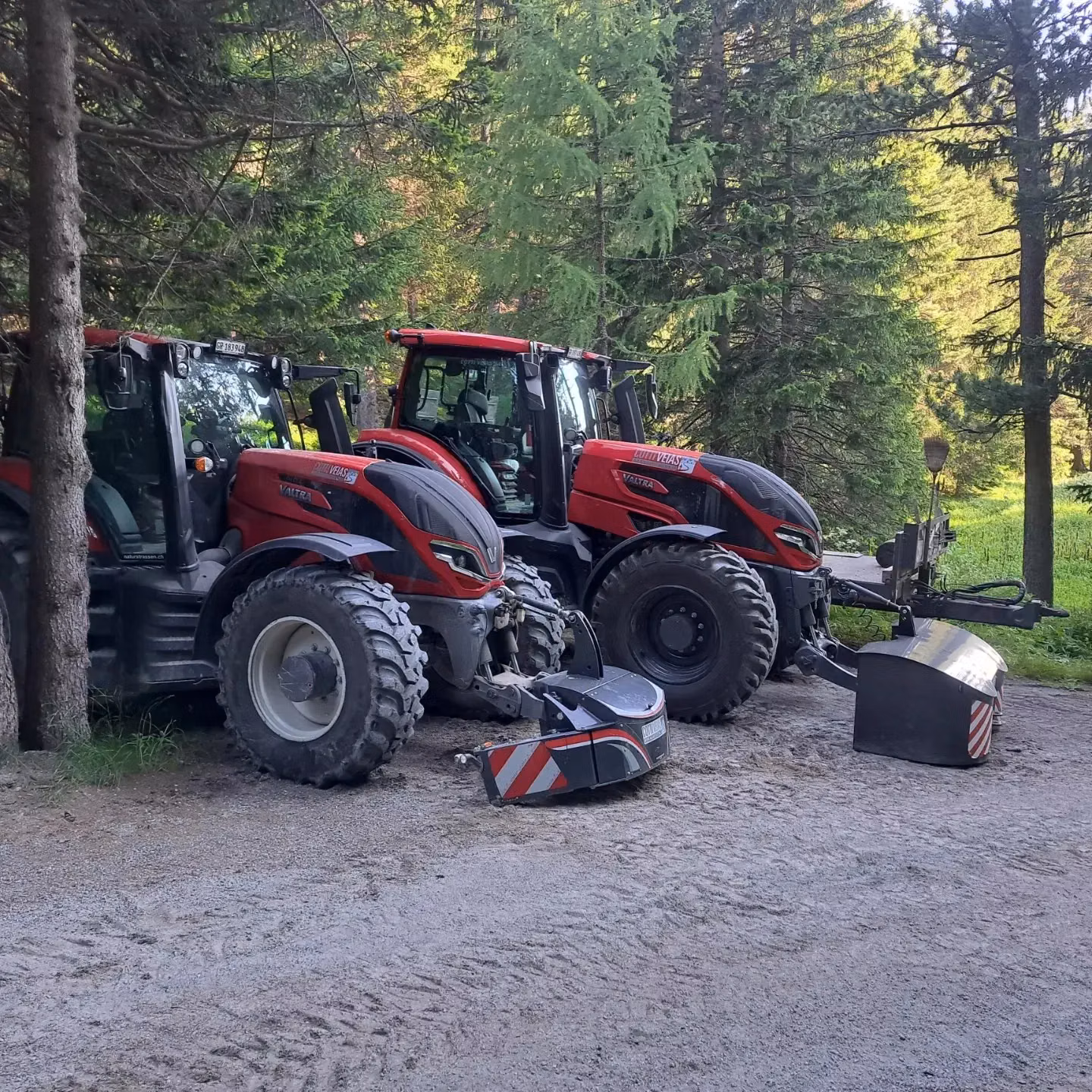Two red tractors are parked in a forest clearing with two detached front weight bumpers on the ground nearby