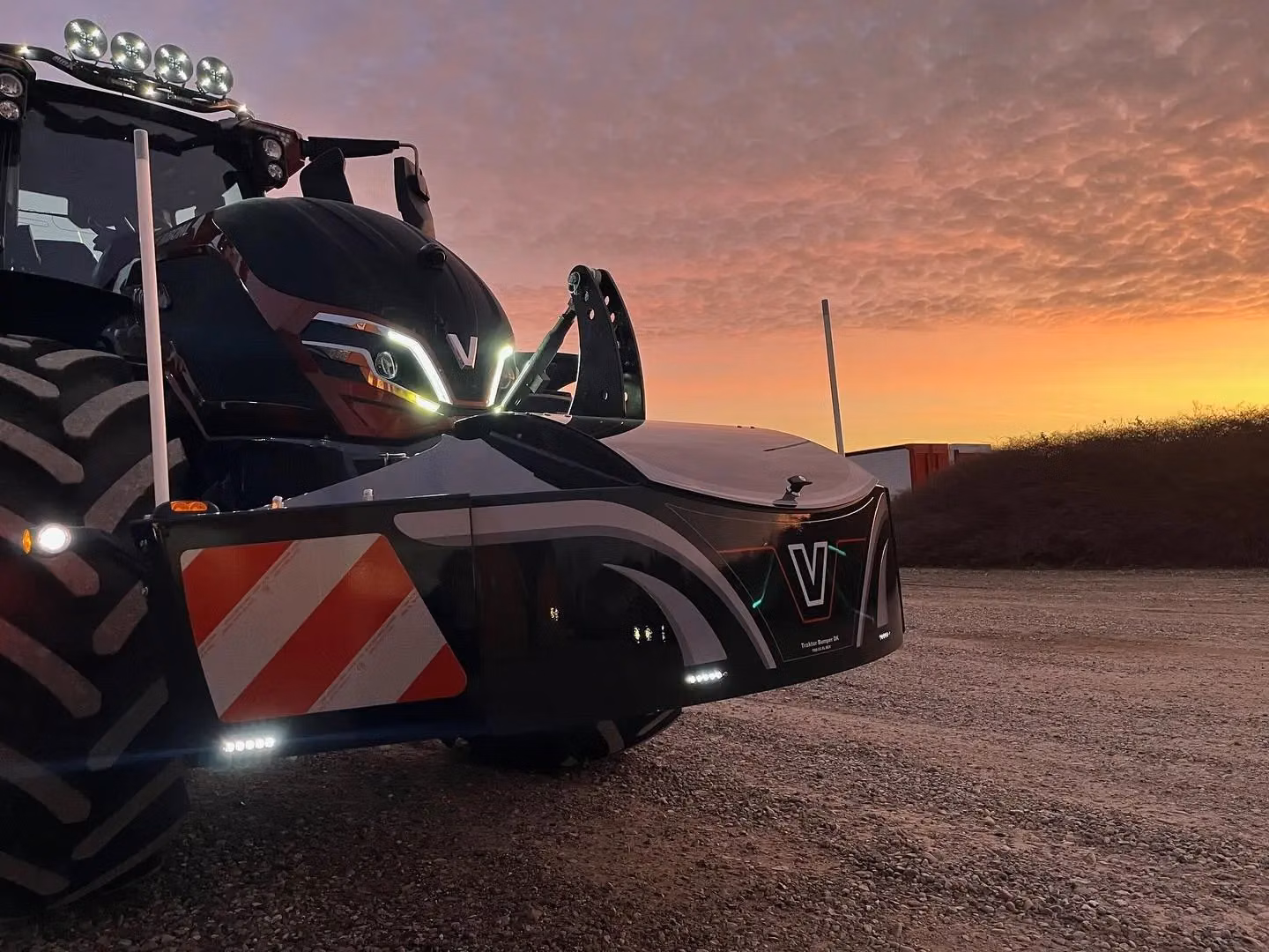 Tractor with large tires and illuminated front weight bumper featuring reflective stripes and V branding at sunset