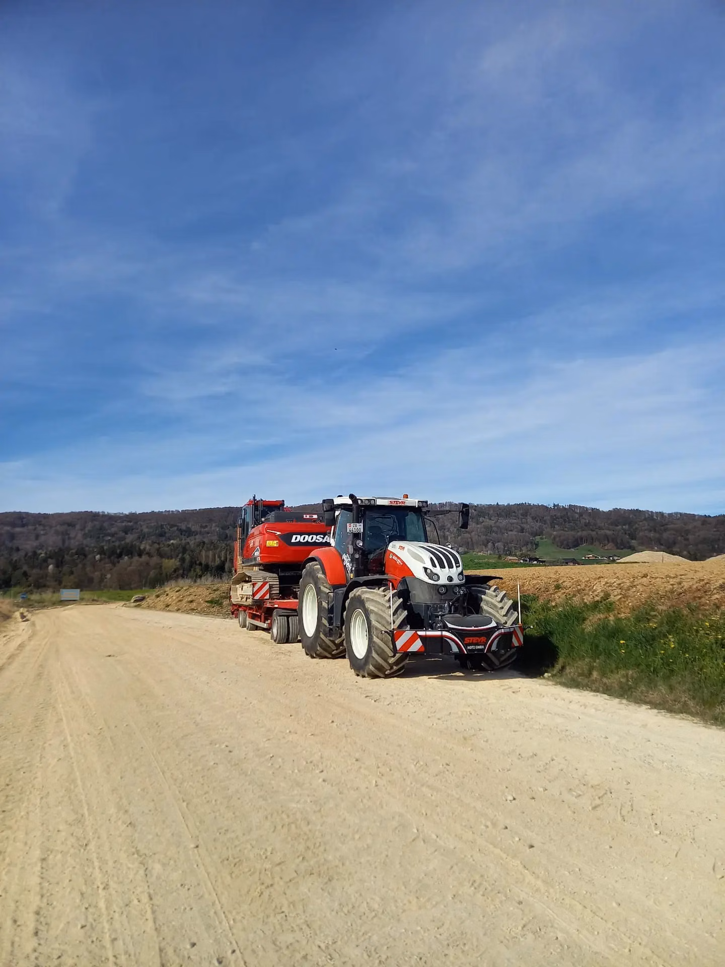 Red and white tractor with a front weight bumper and trailer parked on a dirt road beside grassy fields