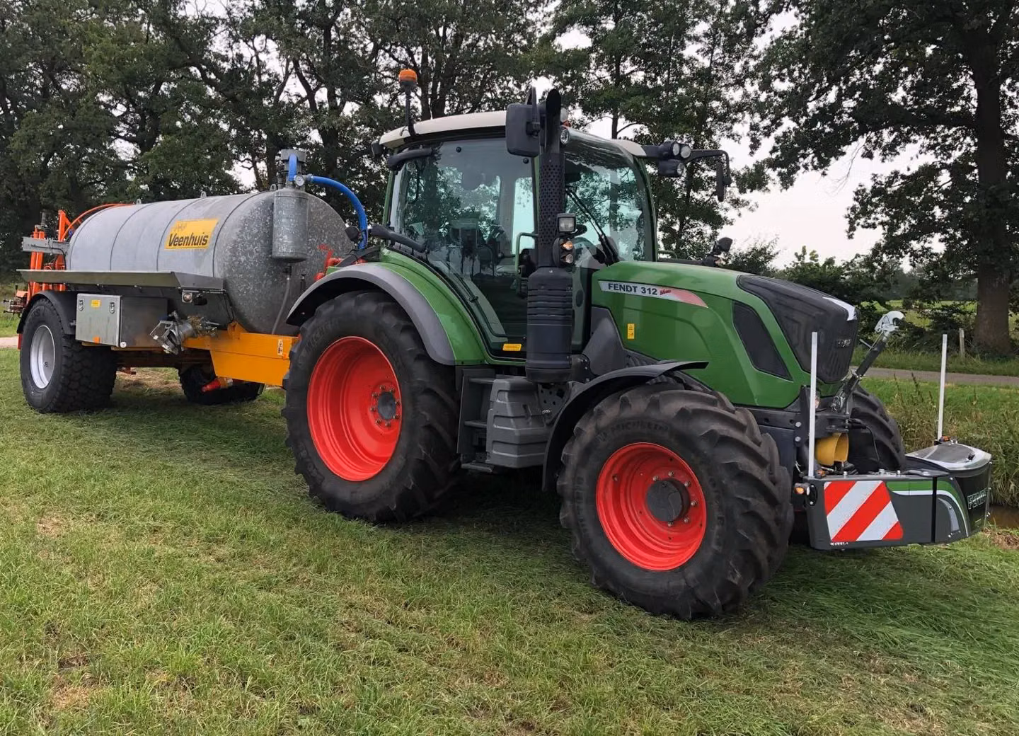 Green tractor with red wheels and front weight bumper attached, pulling a large silver tank trailer on grass