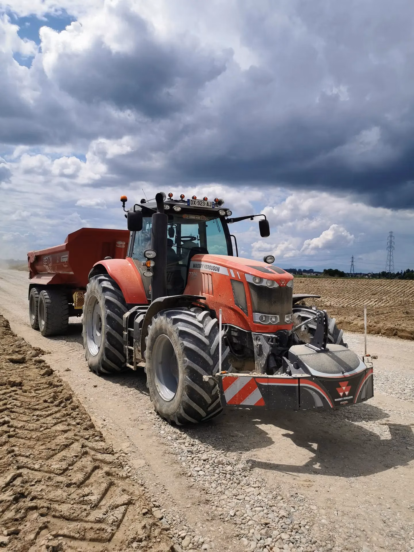 A red tractor with a Massey Ferguson front weight bumper and cargo box is parked on a gravel road under a cloudy sky