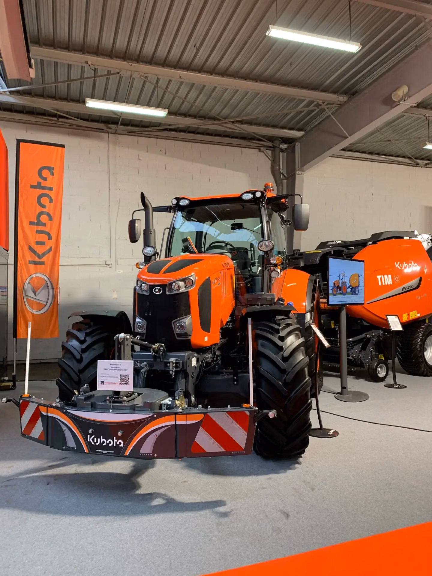 Orange tractor on display indoors with a large front weight bumper labeled Kubota and red-white safety stripes