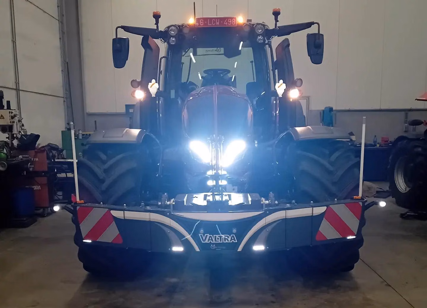 A tractor indoors with headlights on, fitted with a Valtra front weight bumper featuring reflective stripes.