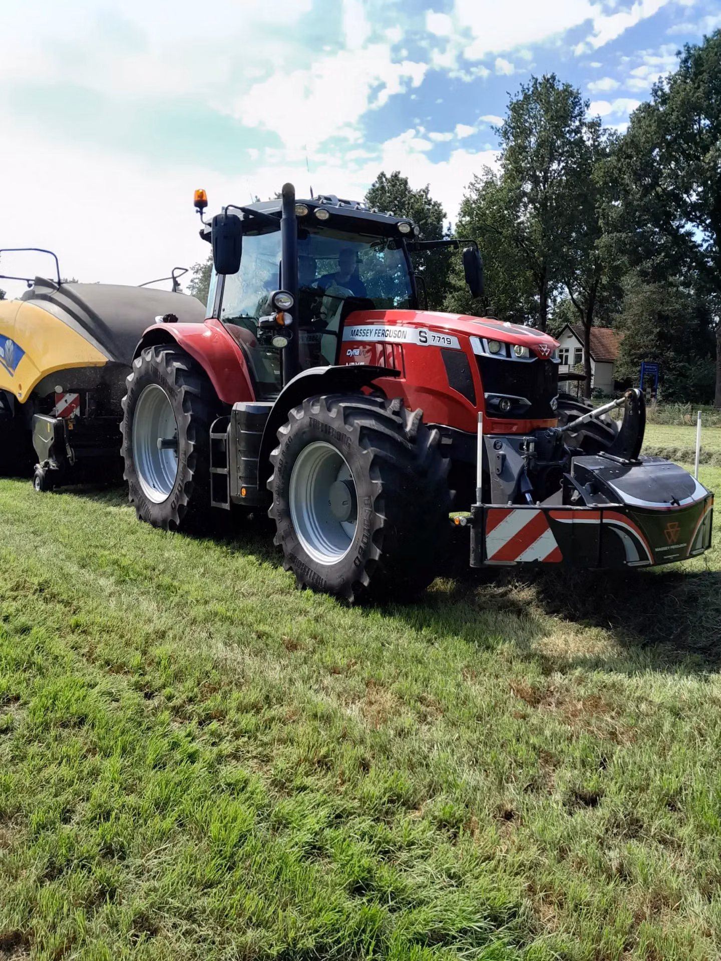 A red tractor on grass with a large front weight bumper attached, pulling yellow agricultural machinery behind