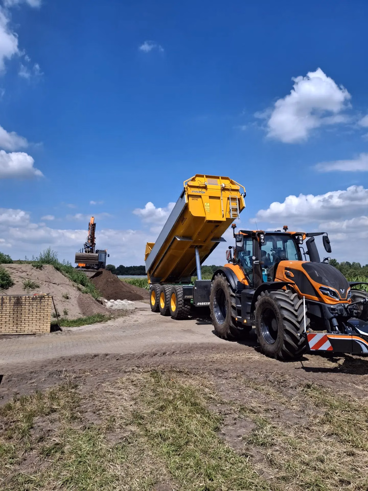 A tractor with a front weight bumper is pulling a yellow trailer on a dirt road under a blue sky with clouds