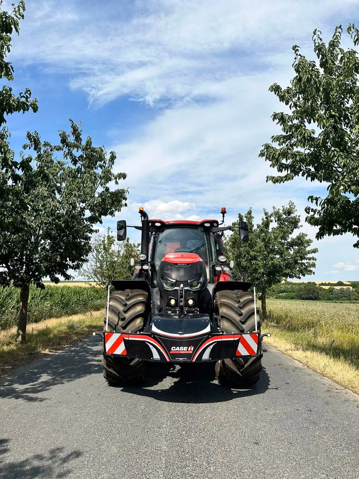 Red tractor with large front weight bumper featuring red and white stripes, parked on a rural road between trees