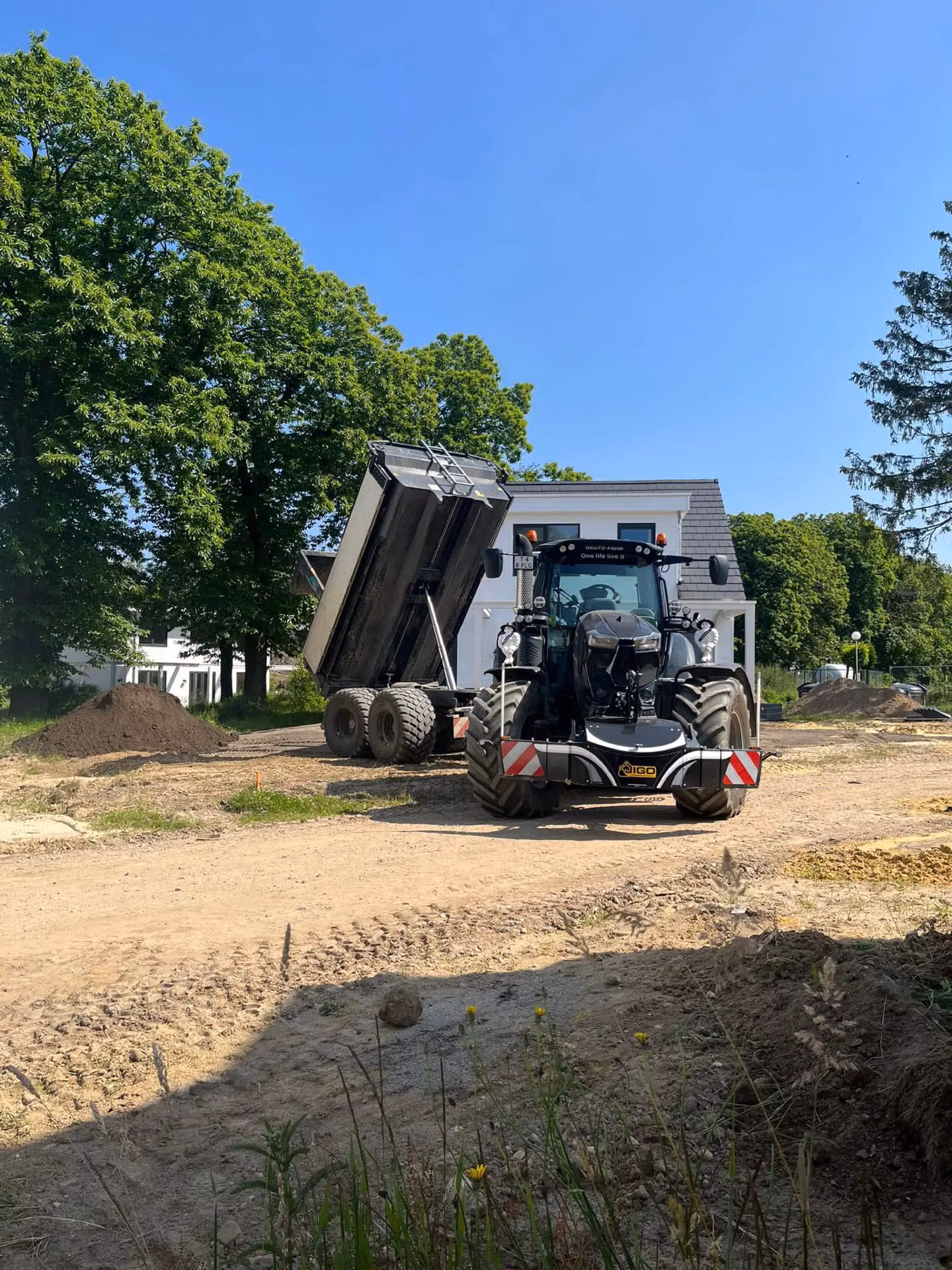 A tractor with a front weight bumper is parked on a construction site beside a trailer with its bed raised