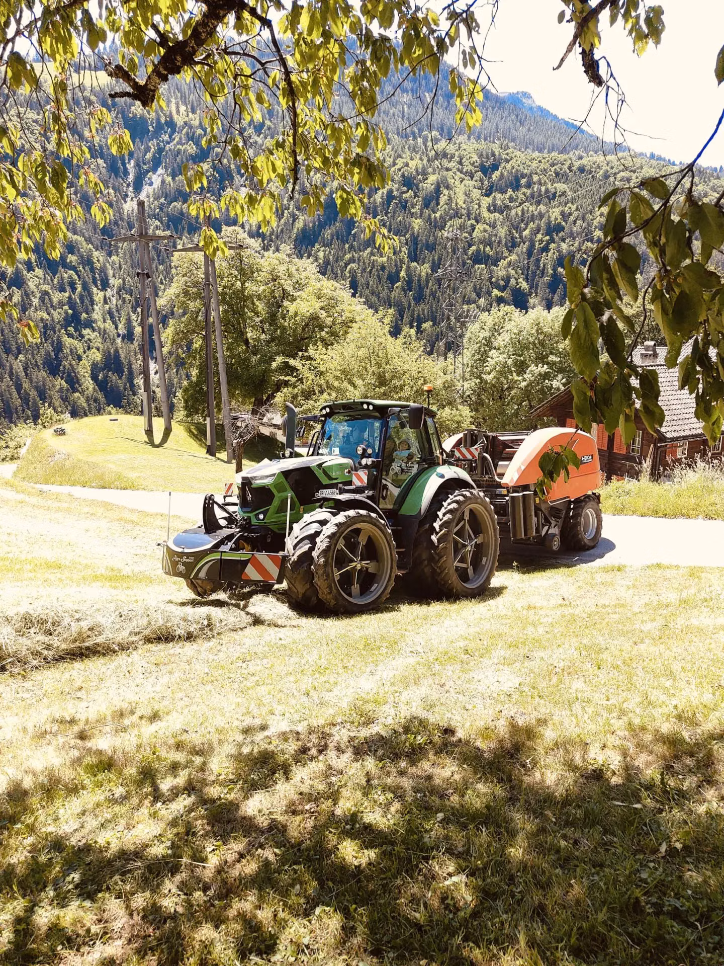 Green tractor with a front weight bumper attached, working on a grassy hillside with trees and mountains in the background