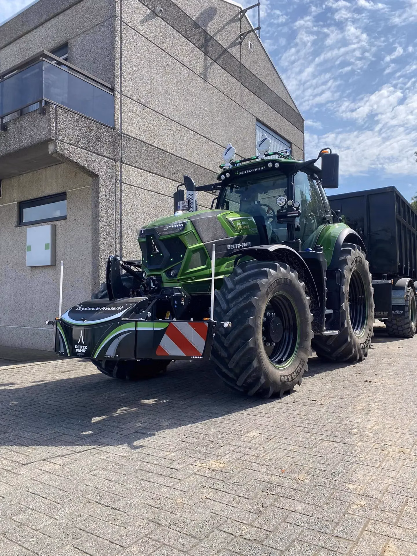 A green tractor with a large front weight bumper attached, parked on paving stones beside a modern building under a blue sky