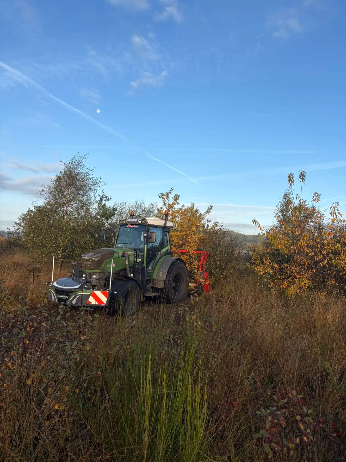 Green tractor with front weight bumper and rear attachment parked in tall grass with trees and blue sky in the background