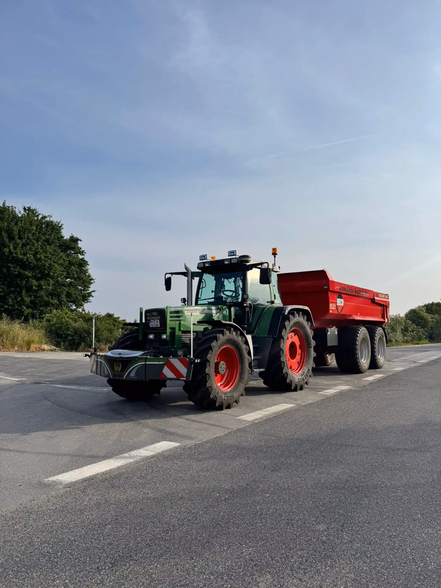 Green tractor with a front weight bumper attached, pulling a large red trailer on an asphalt road