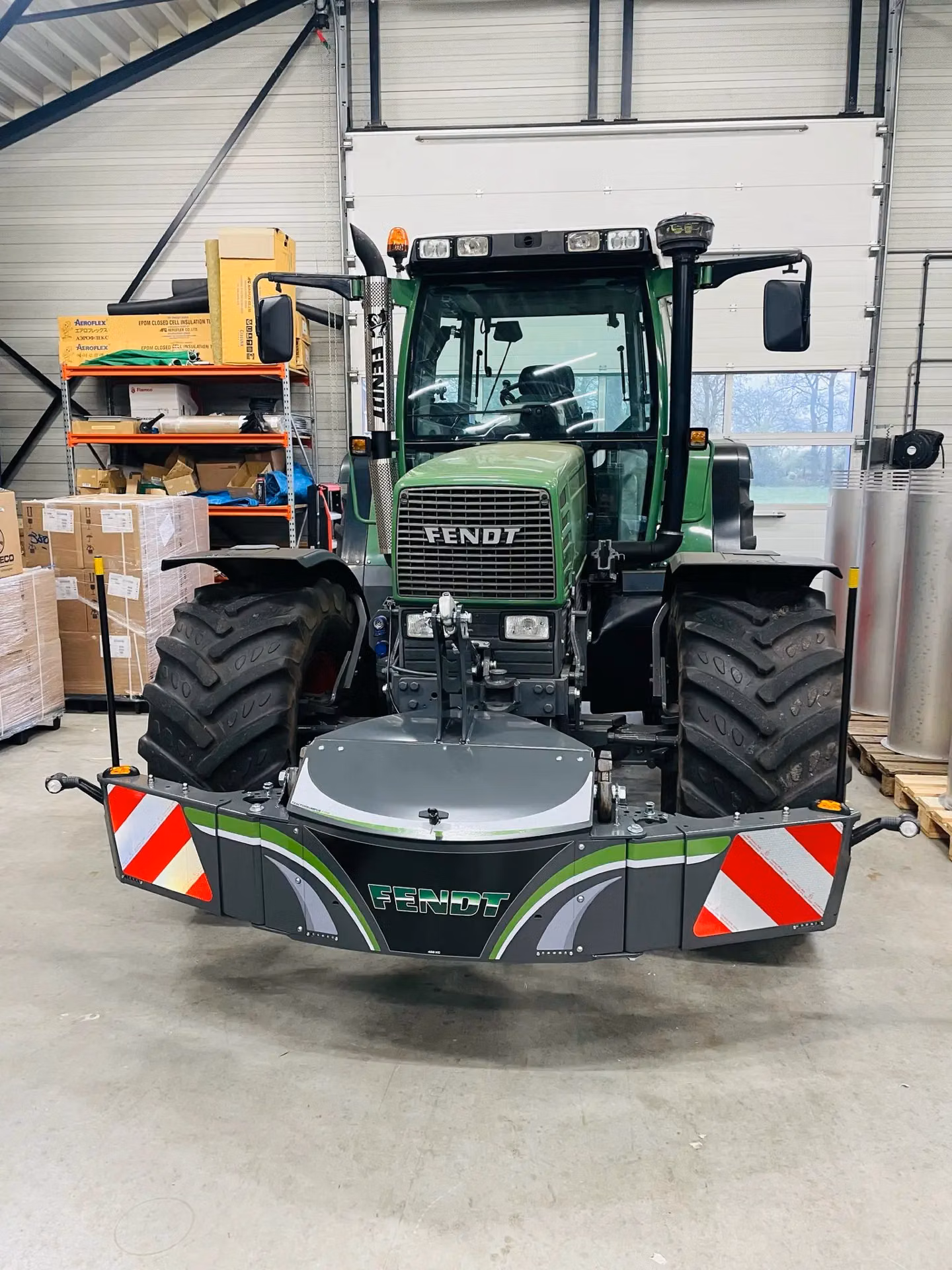 A green tractor with a large front weight bumper, red and white safety reflectors, and Fendt branding in a workshop