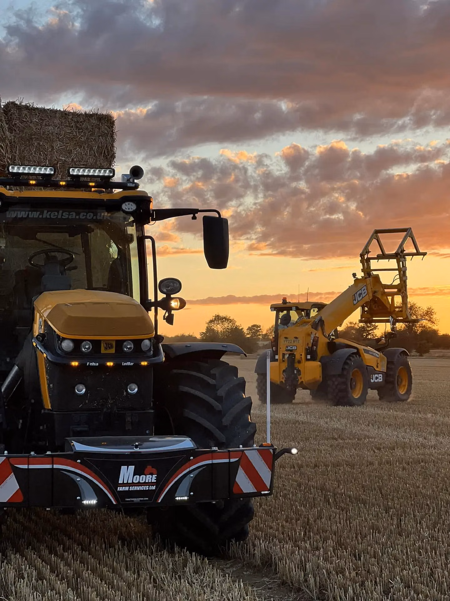 Front view of a tractor in a field at sunset with a front weight bumper and LED lights, carrying hay bales.