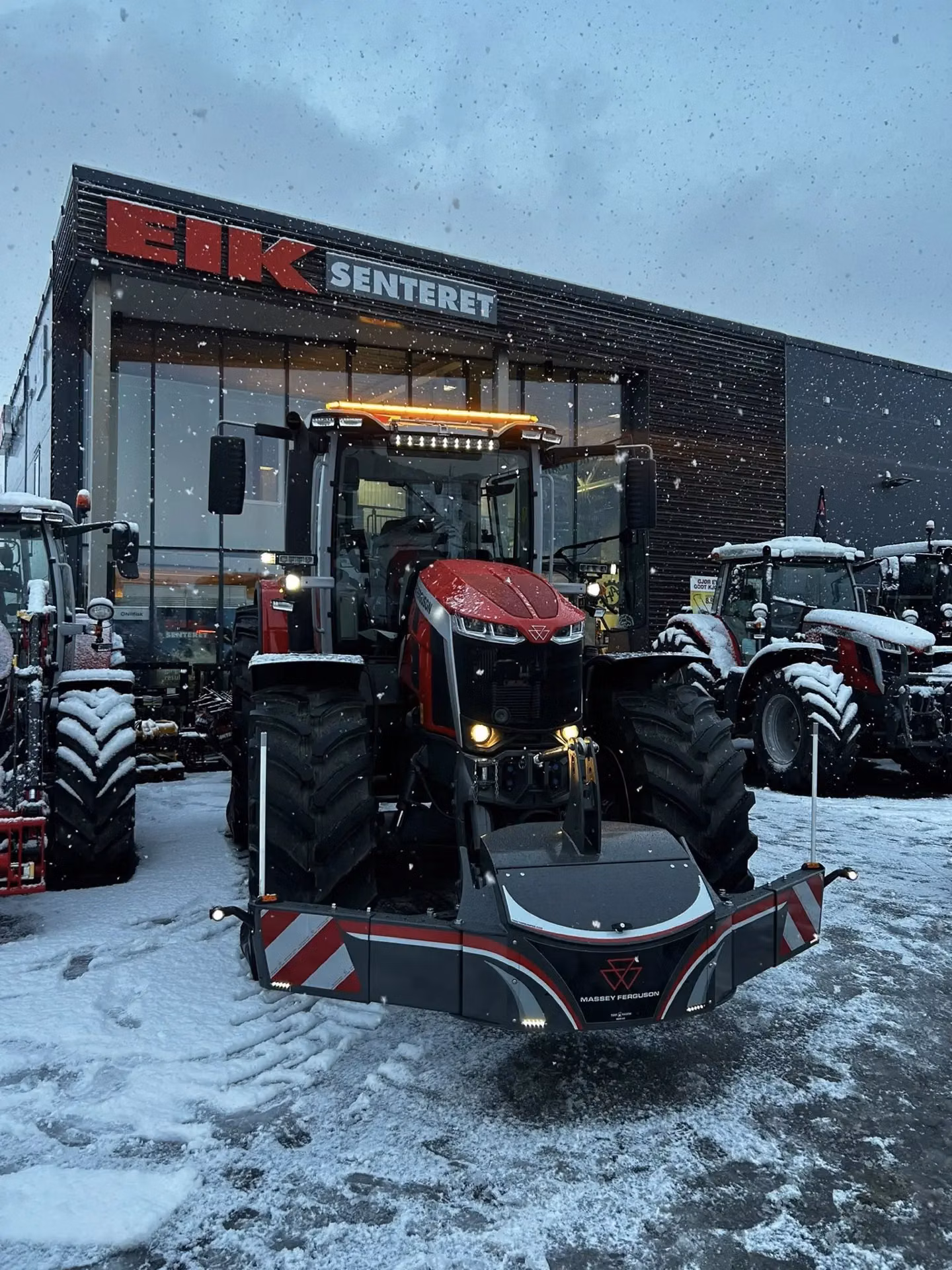 A red Massey Ferguson tractor with a front weight bumper is parked outside a snowy dealership building.