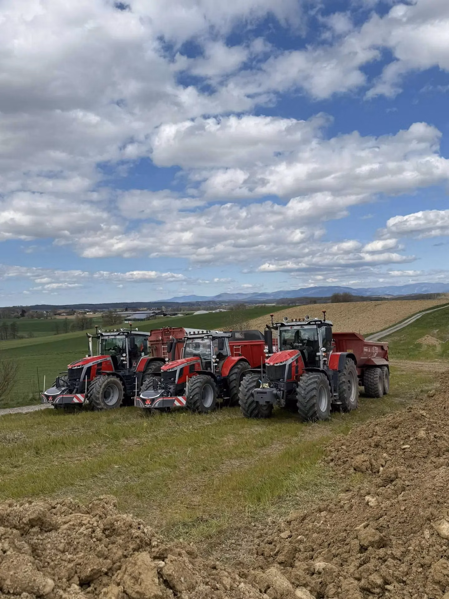 Four red tractors with front weight bumpers and trailers parked on a grassy field under a cloudy sky