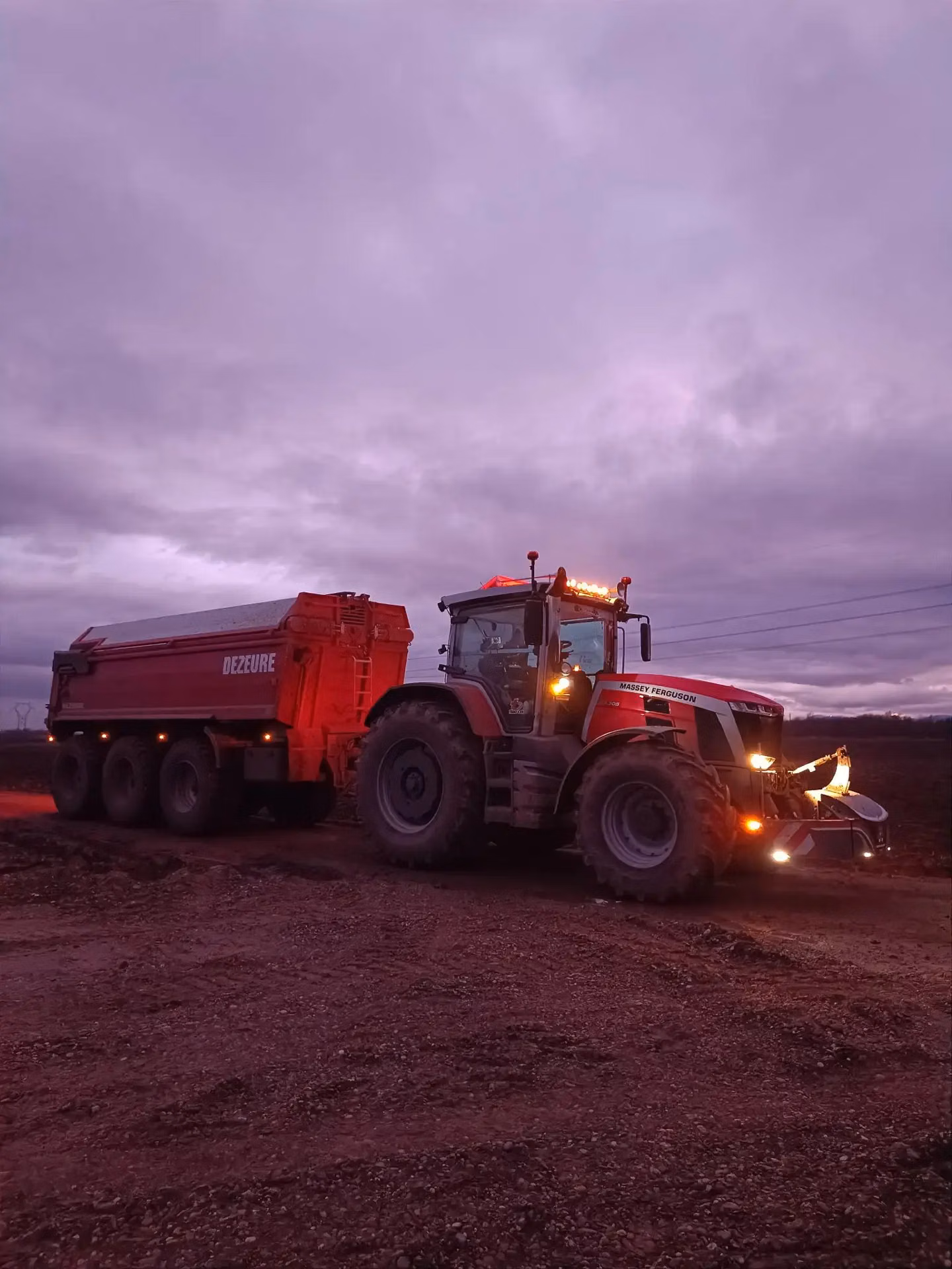 A red tractor with a front weight bumper and trailer attachment is on a dirt road at dusk with lights on
