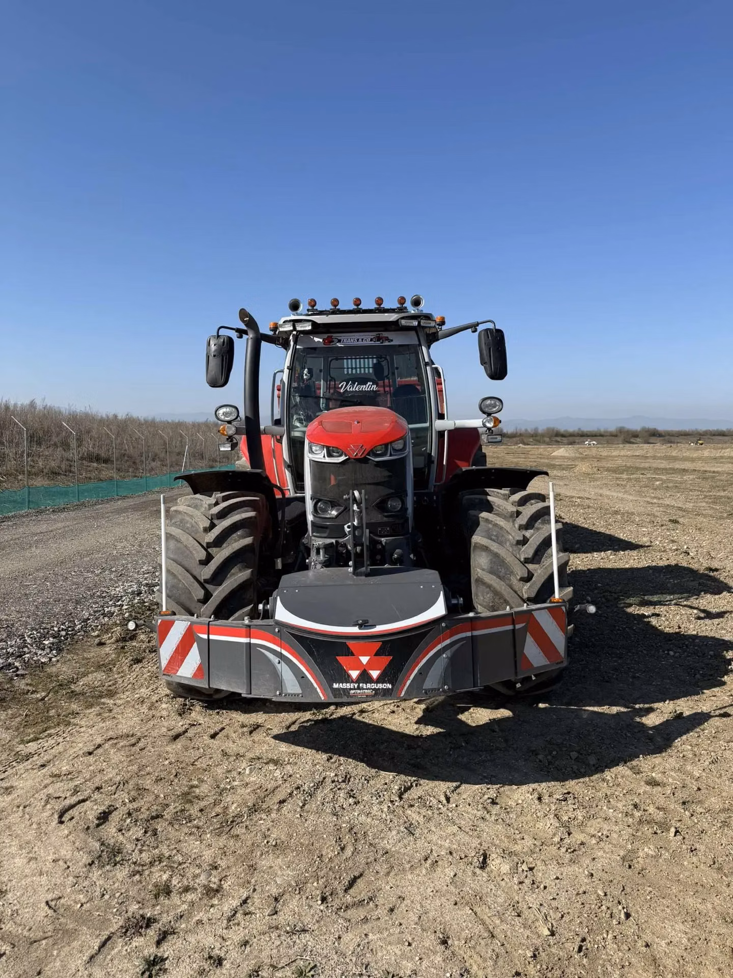 A red tractor with a Massey Ferguson branded front weight bumper is parked on a dirt field under a clear sky