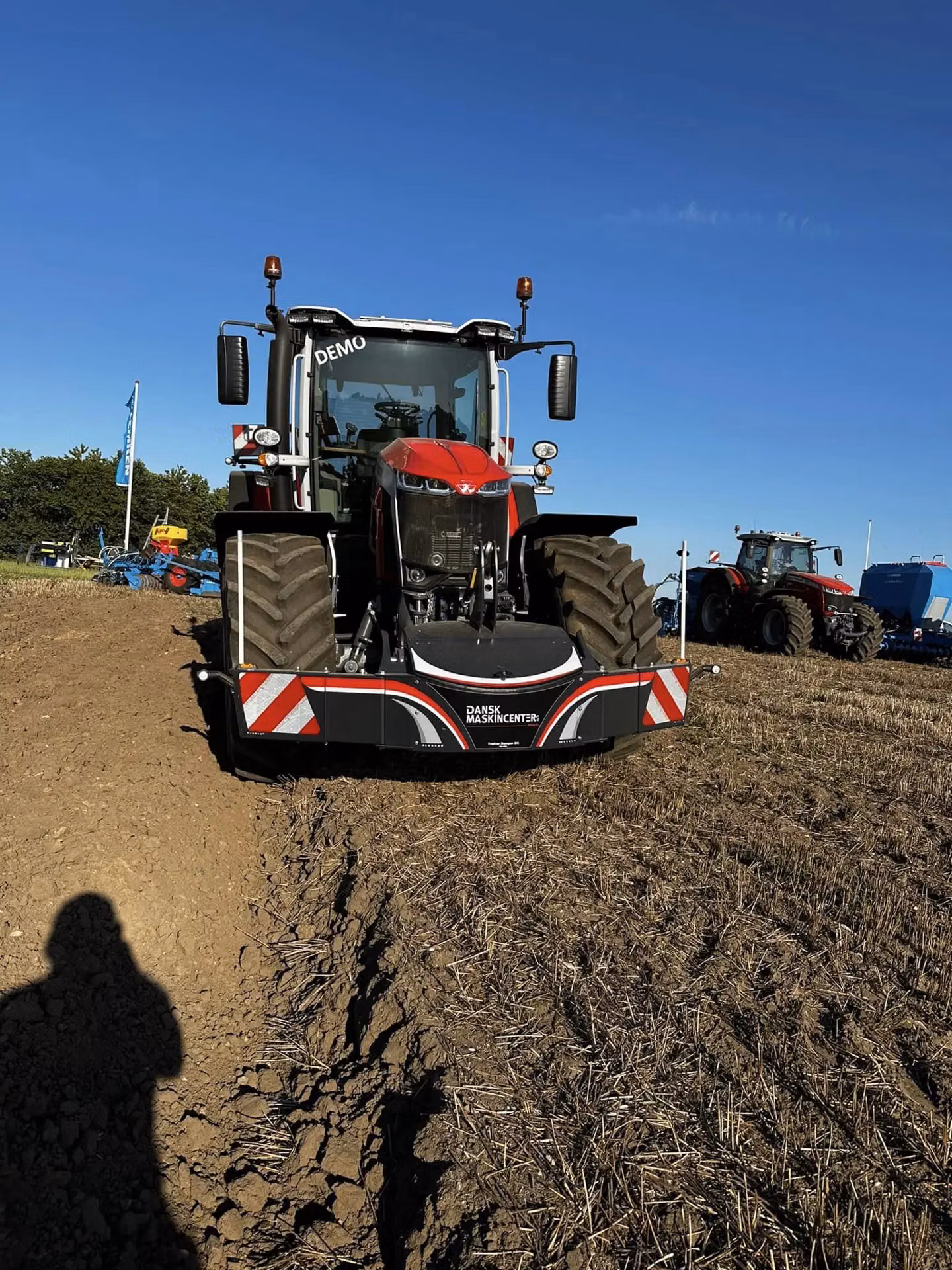 A red tractor with a large front weight bumper is parked on a cultivated field under a clear blue sky