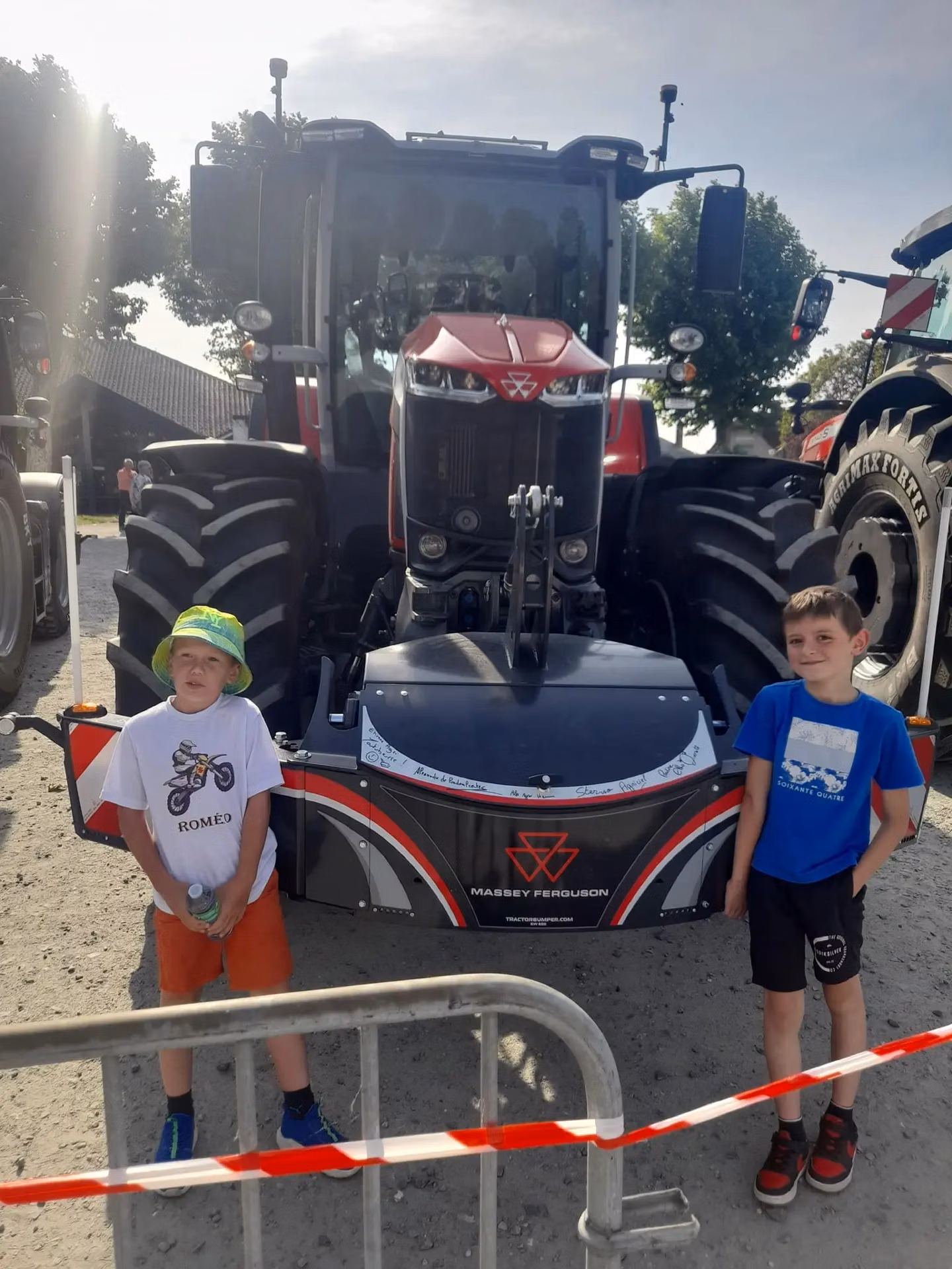 Two boys stand in front of a Massey Ferguson tractor with a front weight bumper behind a barrier and tape