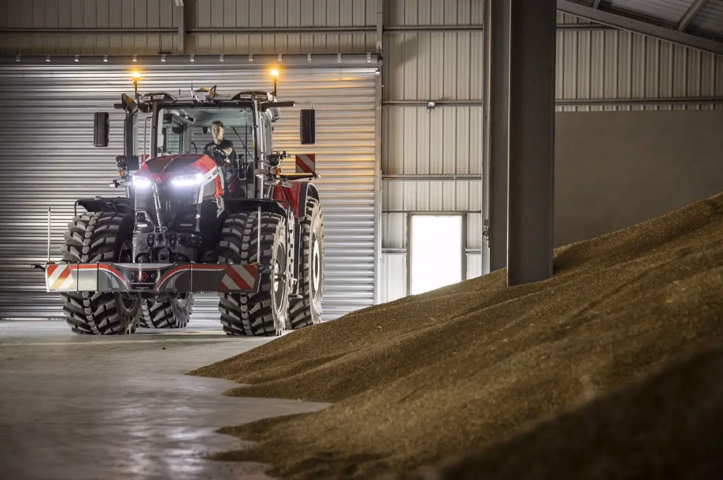 Red tractor with a front weight bumper and dual wheels parked indoors near a large pile of grain