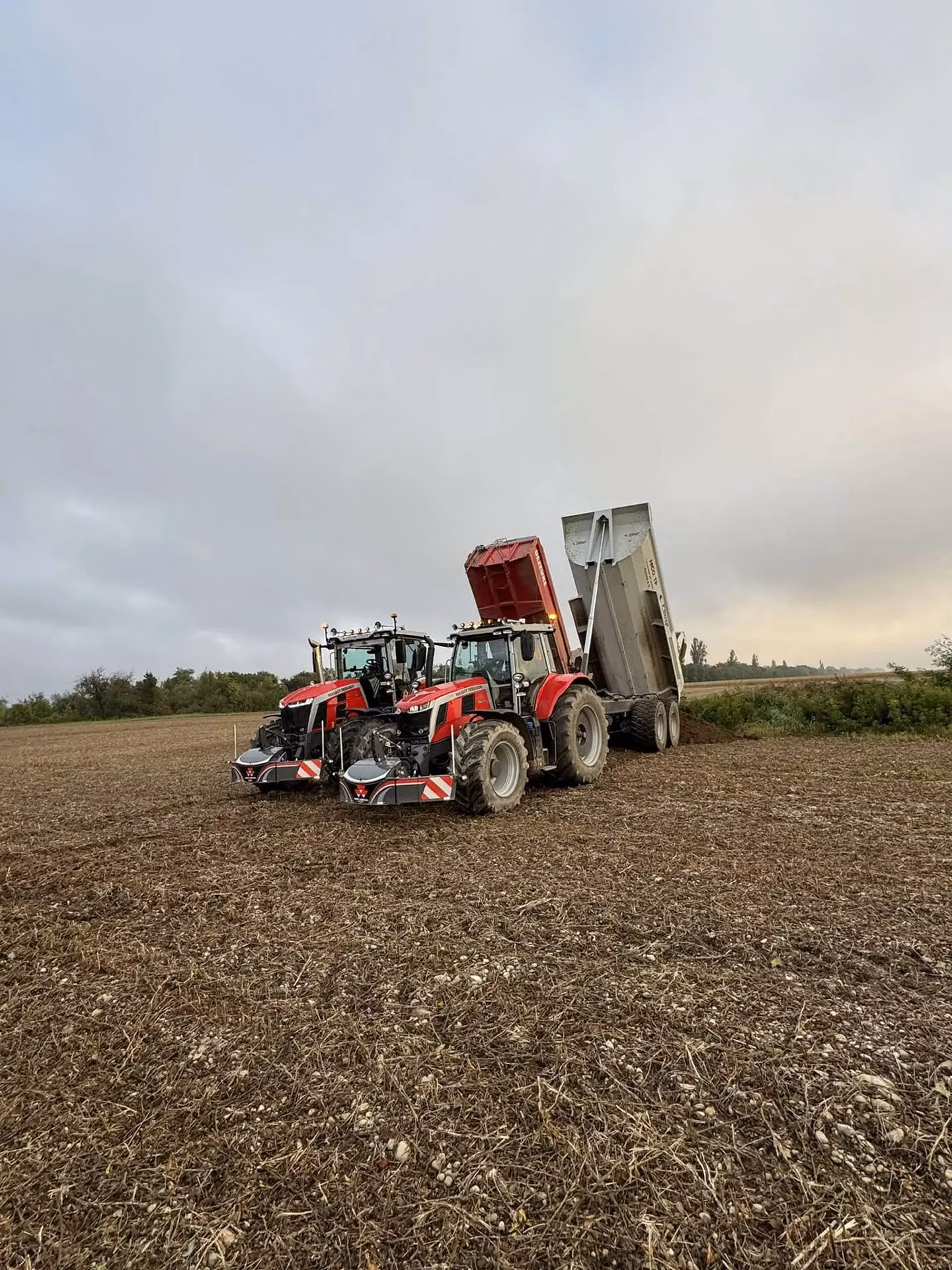 Two red tractors with front weight bumpers are parked in a field next to a raised trailer.
