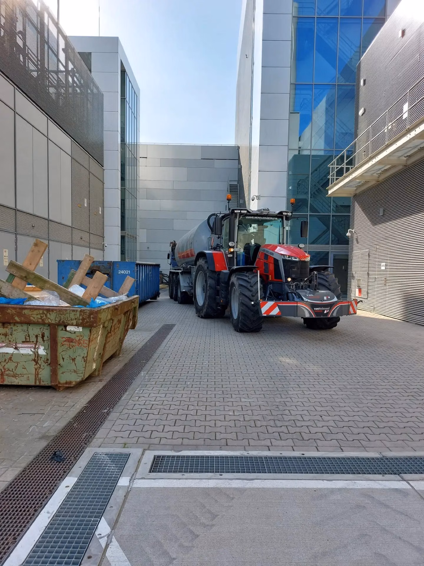 A red tractor with a front weight bumper and cargobox is parked between modern industrial buildings