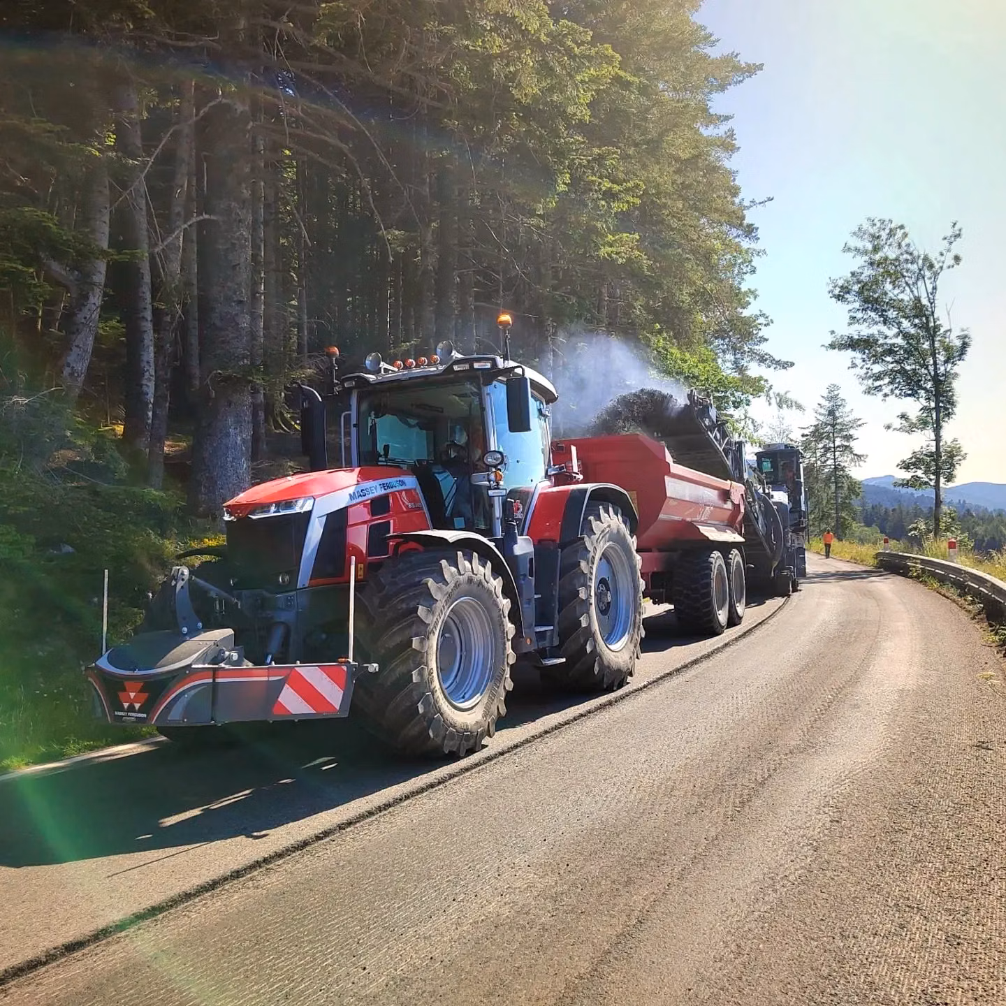 A red tractor with a front weight bumper is parked on a forest road, attached to a large trailer being filled with material