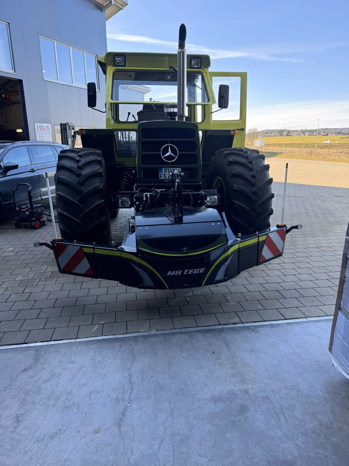Green tractor with large front tires fitted with an MB trac front weight bumper on a paved area outside a building