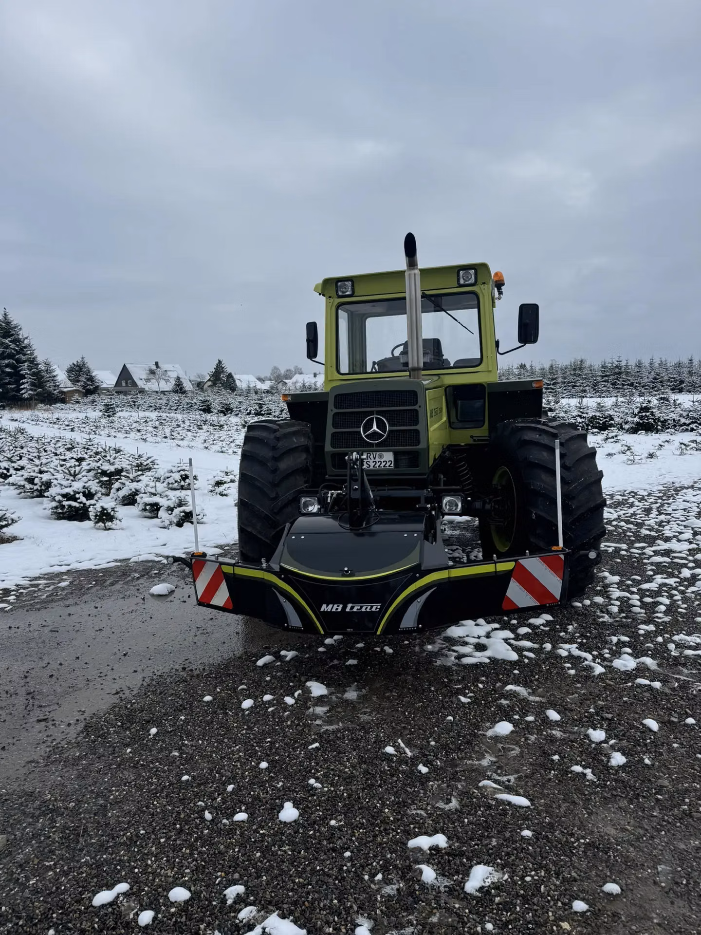 Green tractor with large tires and an MB trac front weight bumper, parked on a snowy gravel surface
