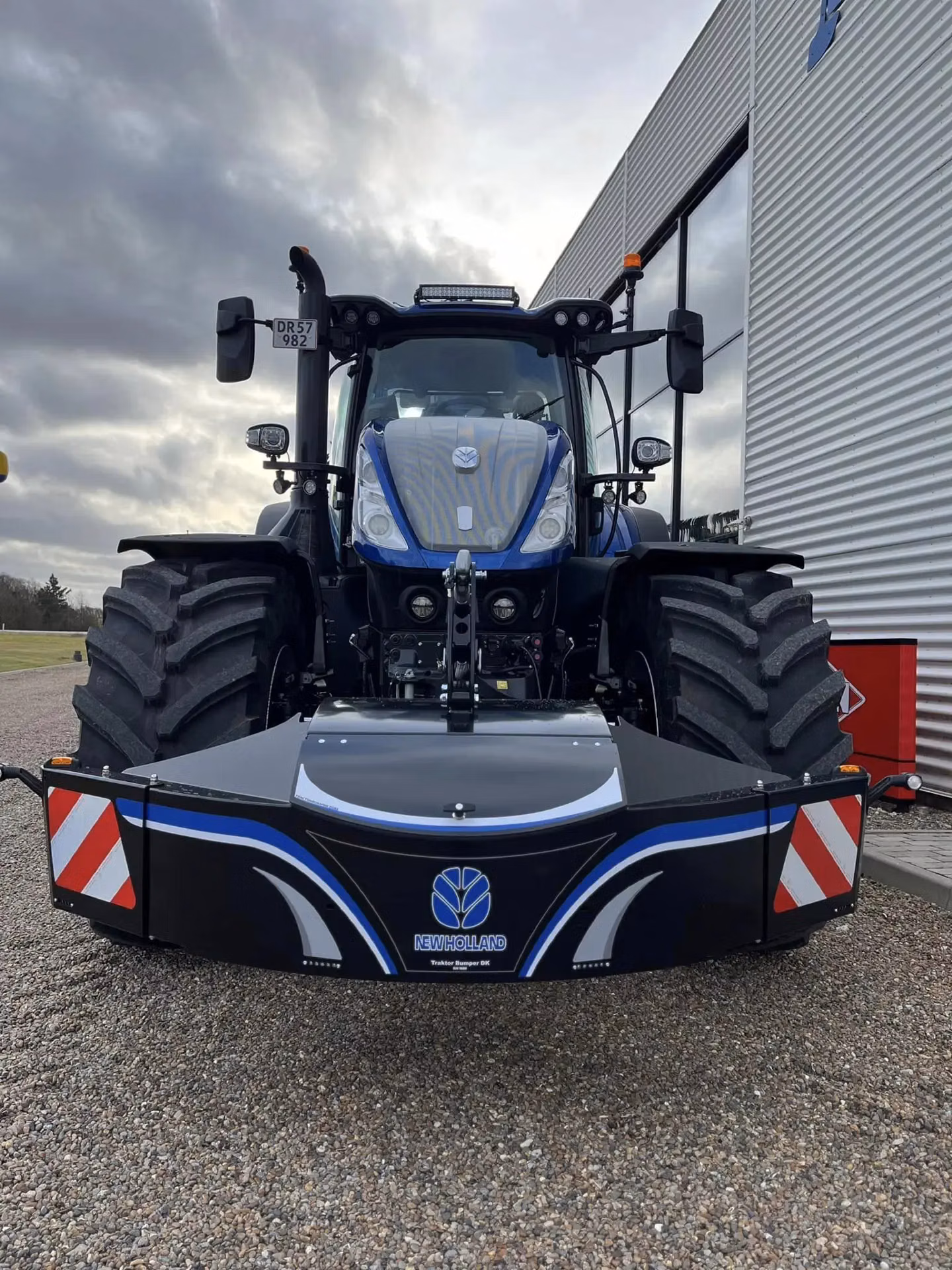 Large blue tractor with a New Holland branded front weight bumper and reflective safety markings on gravel ground