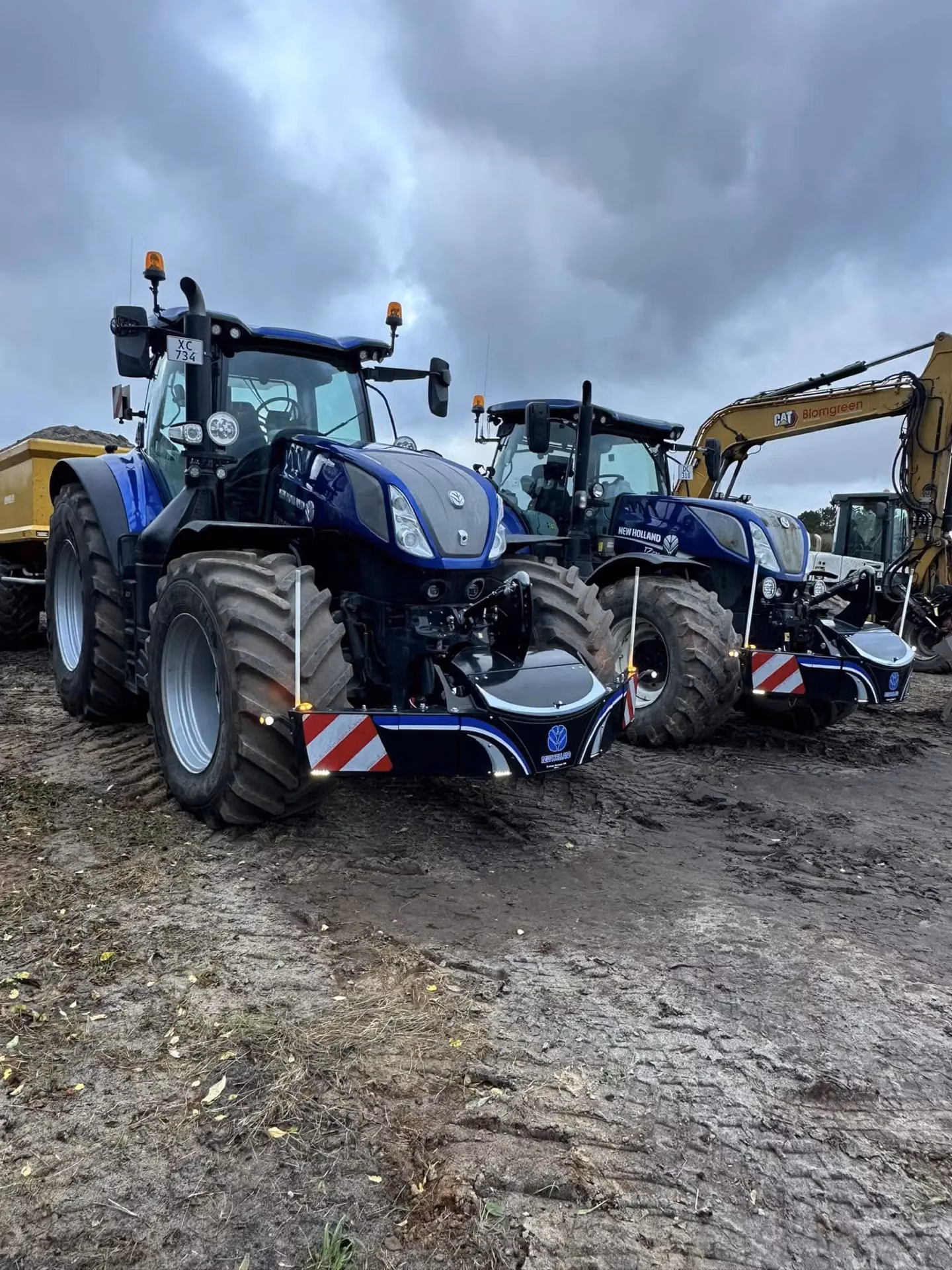 Two blue tractors with front weight bumpers are parked on muddy ground under a cloudy sky, with machinery nearby