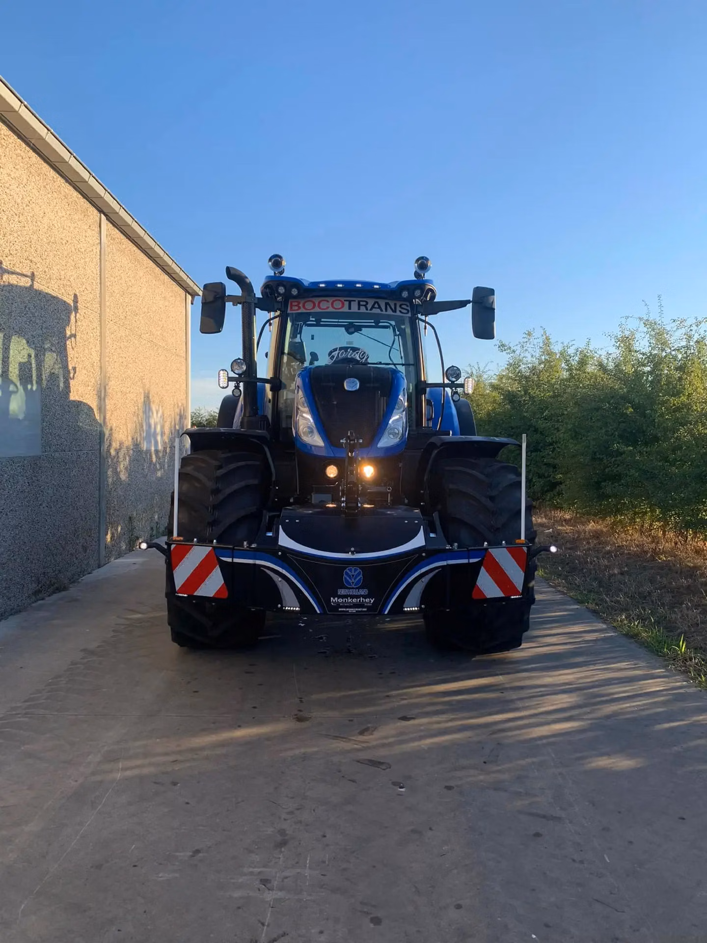 A blue tractor with a mounted front weight bumper featuring reflective red and white stripes on a concrete path