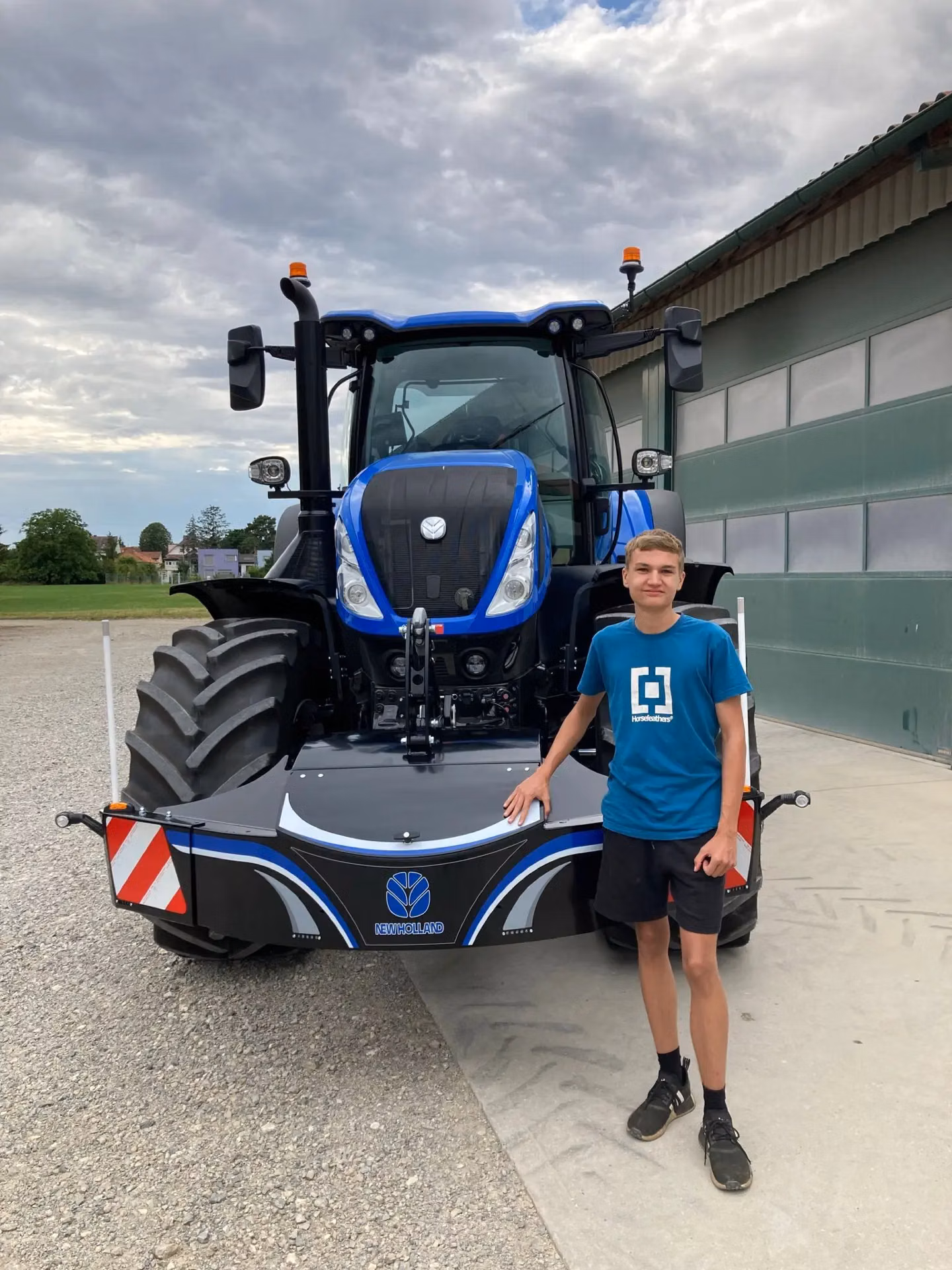 A person stands beside a blue tractor fitted with a New Holland branded front weight bumper with LED lights.