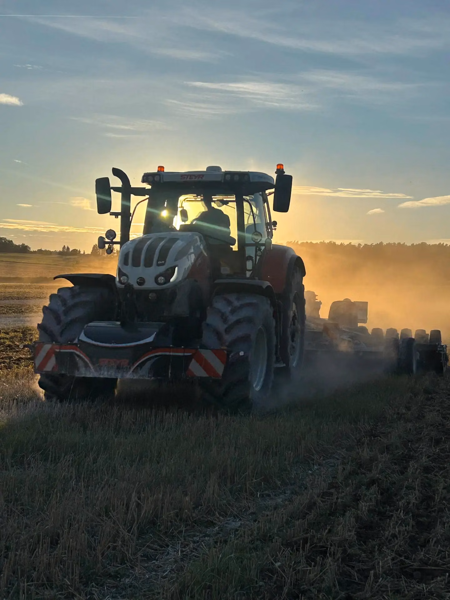 Tractor with a striped front weight bumper working in a field at sunset, kicking up dust as it moves forward