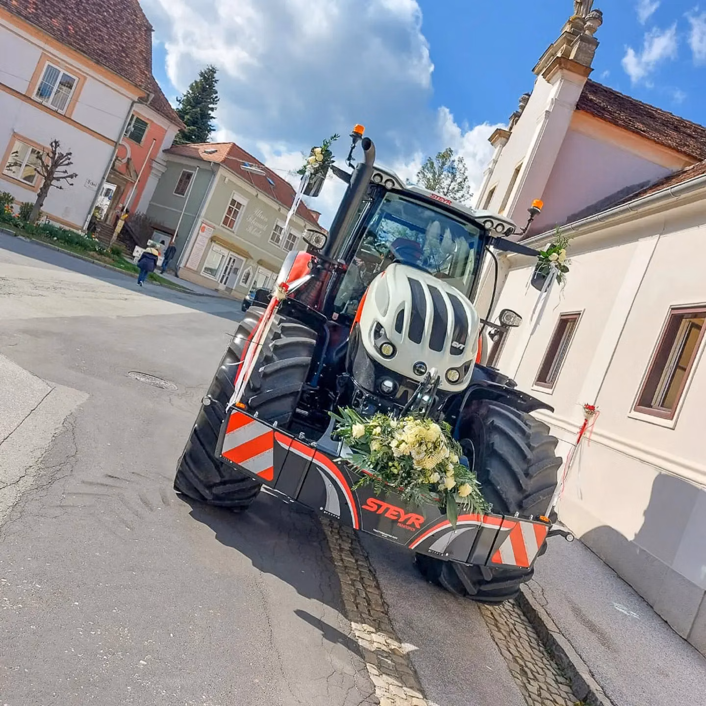 Tractor with a front weight bumper labeled STEYR, decorated with flowers, parked on a street near buildings