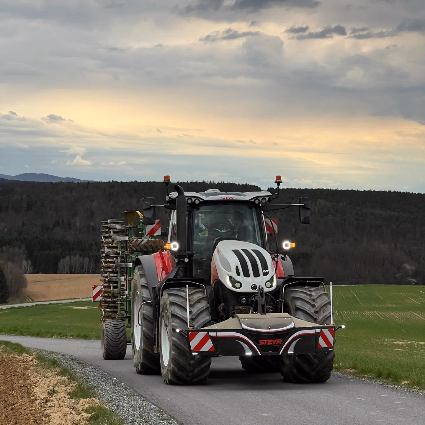 A red and white tractor with a mounted front weight bumper drives on a rural road with fields and forest in the background
