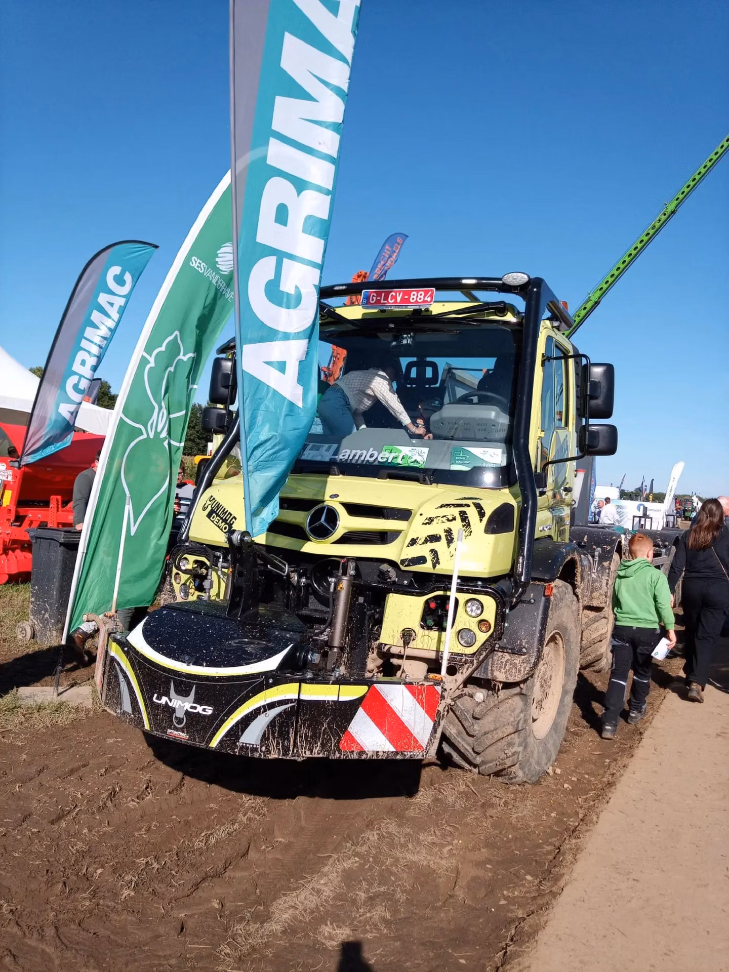 Muddy tractor with a front weight bumper labeled Unimog on display between green AGRIMAC flags at an outdoor event