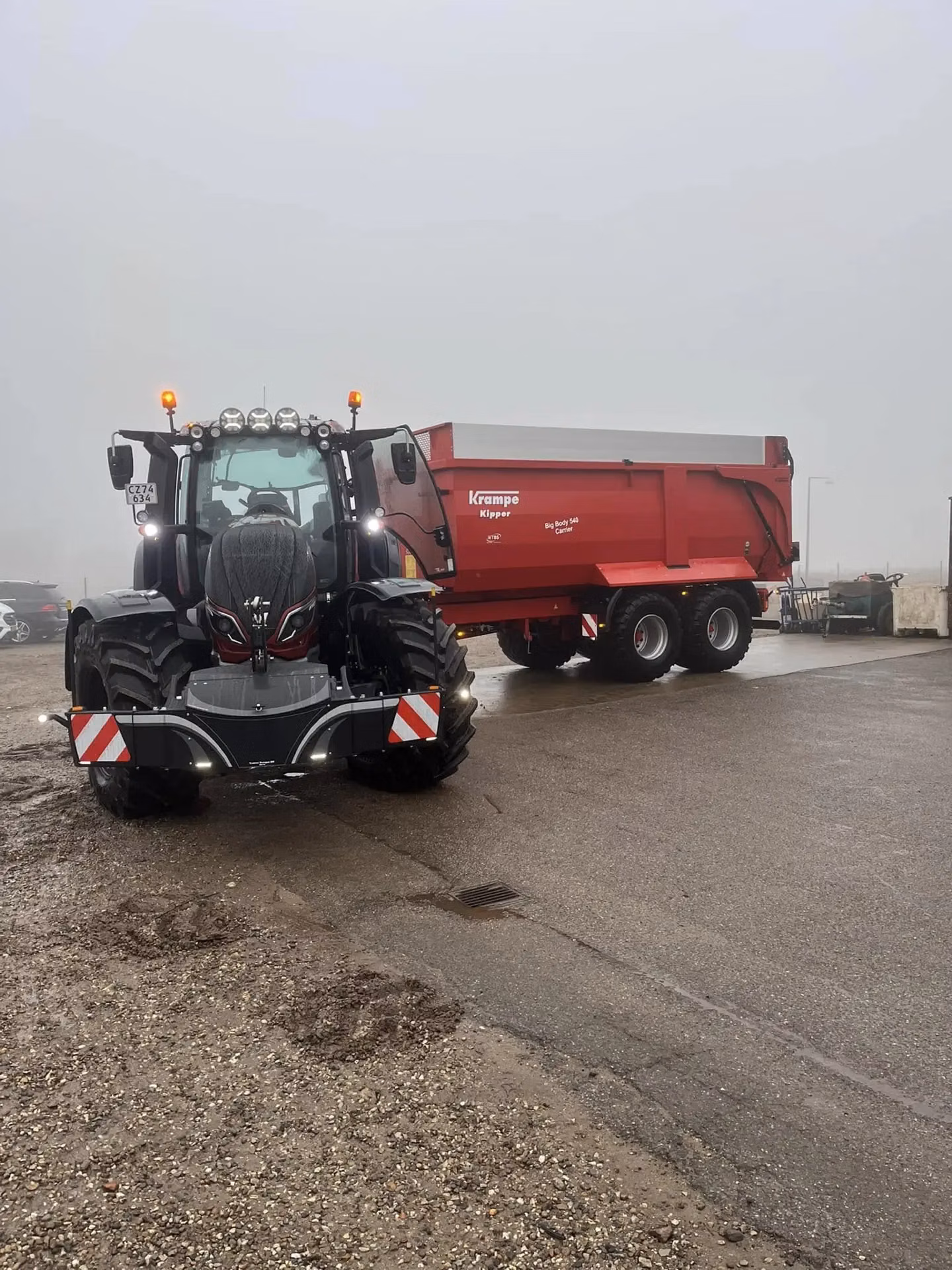 A tractor with a mounted front weight bumper and a red trailer attached, parked on a wet, overcast day