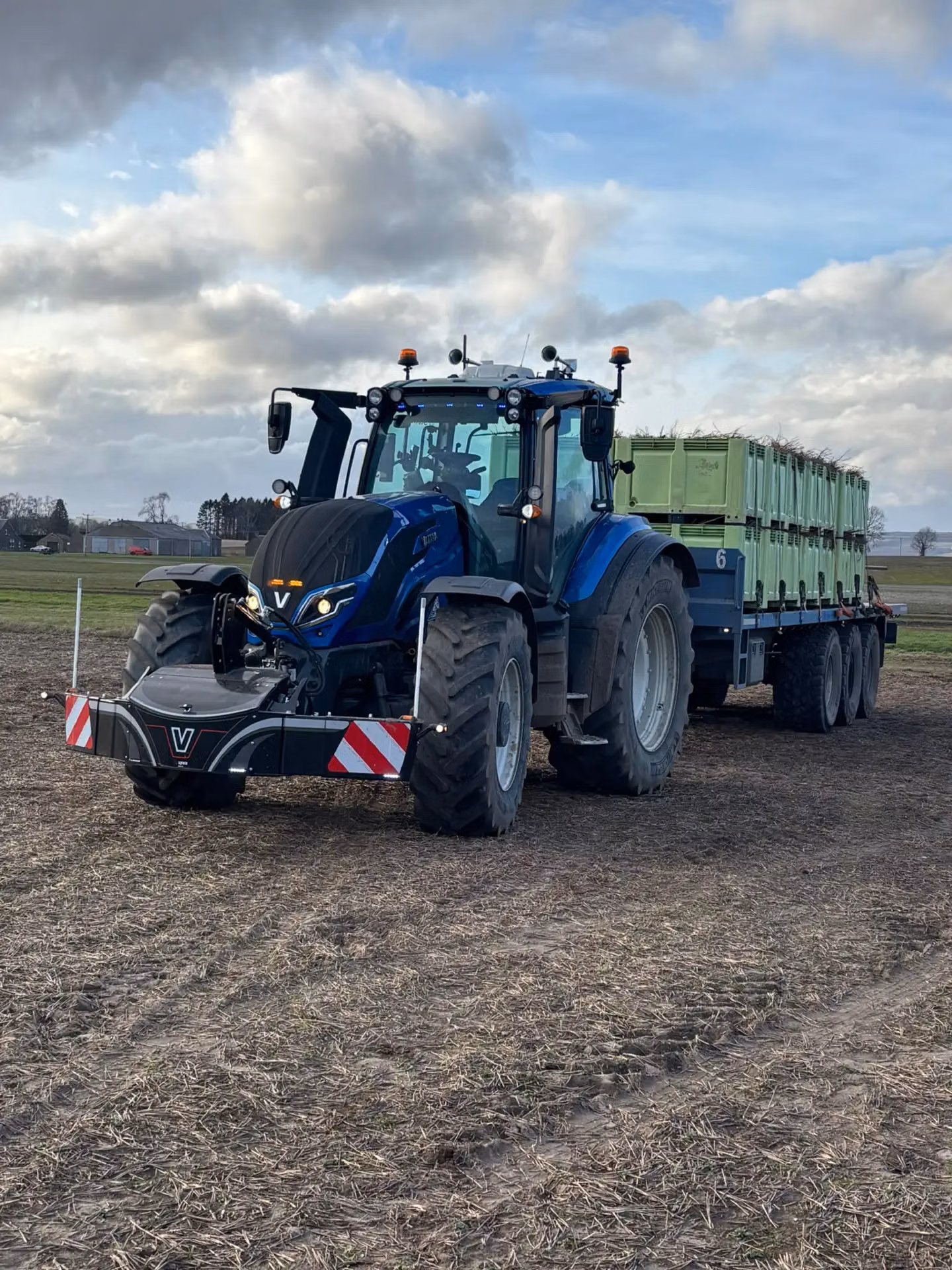A blue tractor with a front weight bumper and warning stripes, towing a trailer loaded with green crates in a field