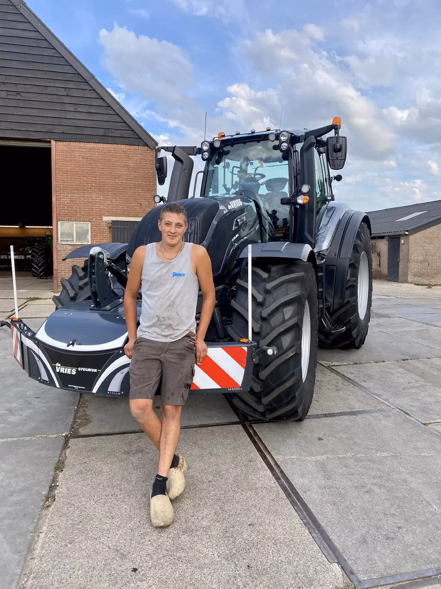 Person in work clothes and wooden clogs stands in front of a black tractor with a front weight bumper and LED lights