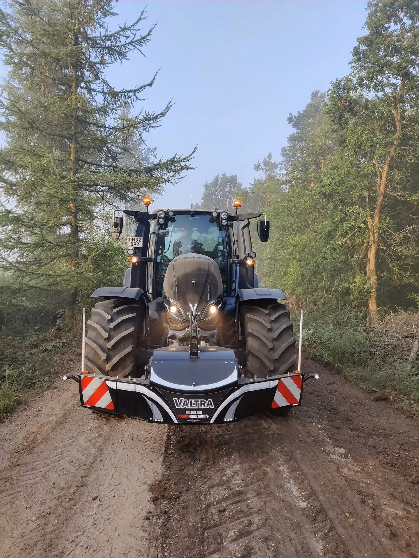 Black tractor with a front weight bumper labeled Valtra on a dirt track surrounded by trees and foggy sky