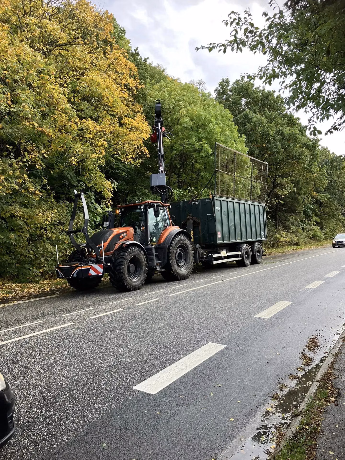 A tractor with a front weight bumper and a mounted crane is parked on a roadside hauling a large green trailer