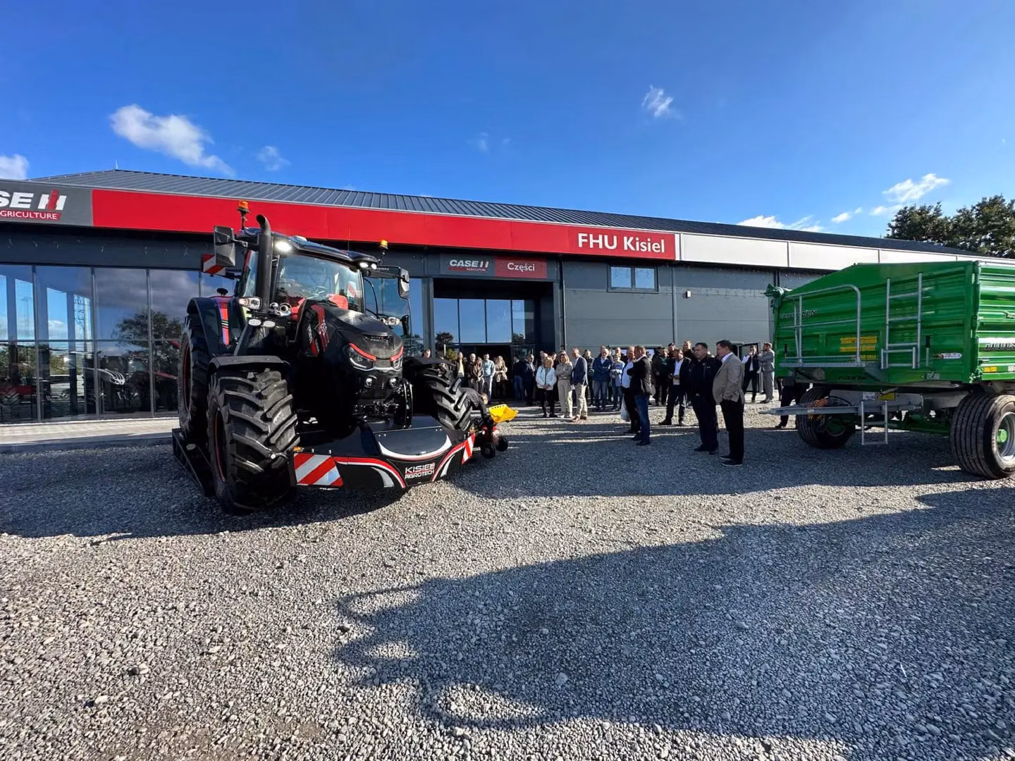 Black tractor with a front weight bumper attached, parked outside a building with people and a green trailer nearby