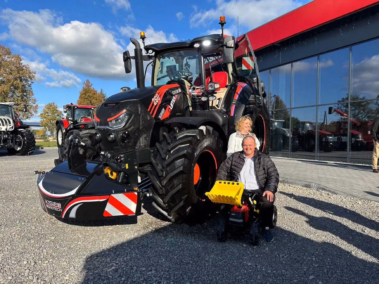 Large black tractor with a front weight bumper attachment and two people posing in front outside a glass building