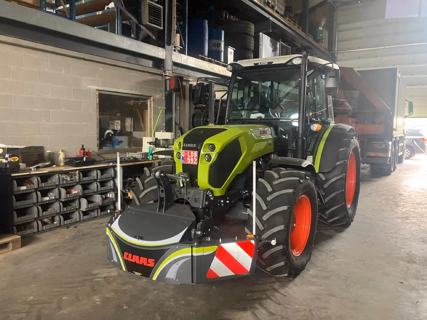 Green tractor with red wheels in a workshop, fitted with a front weight bumper labeled CLAAS and red reflective stripes