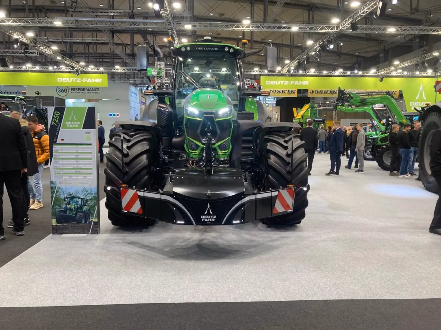 Large green tractor with a black front weight bumper and red-white safety stripes on display at an indoor exhibition